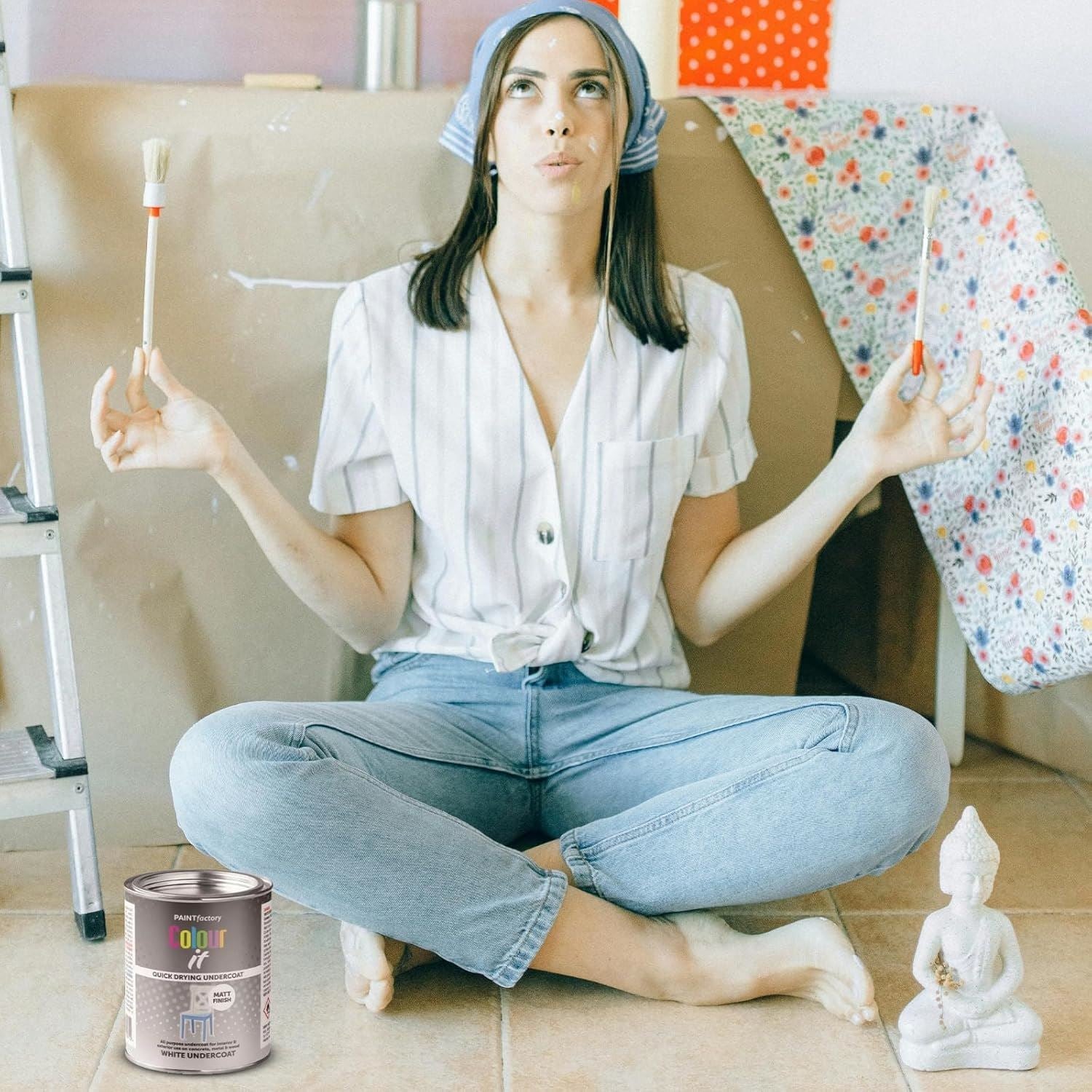 A woman meditating with paintbrushes holding a tin of white matt undercoat paint 300ml on the floor